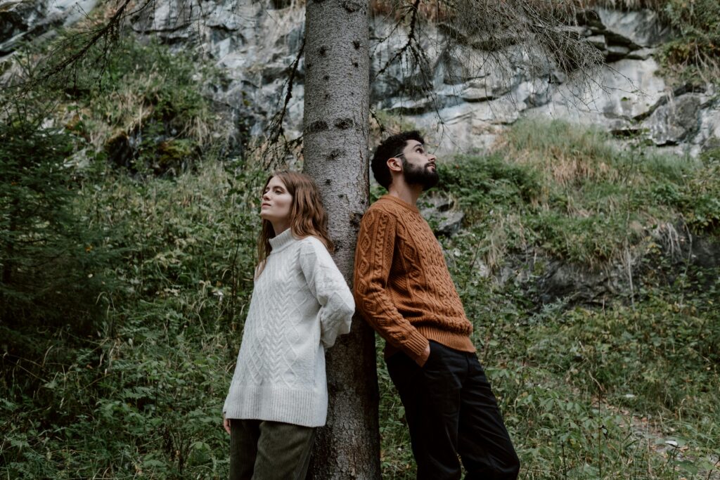 A couple stands back to back against a tree in an autumn forest setting.