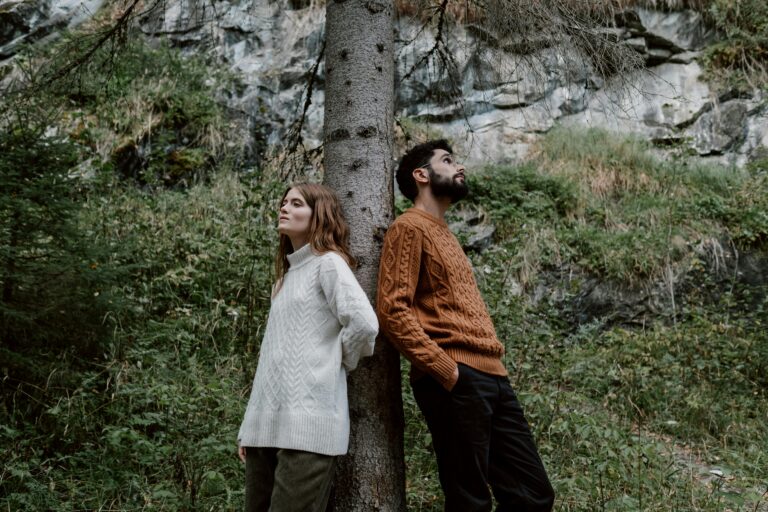 A couple stands back to back against a tree in an autumn forest setting.